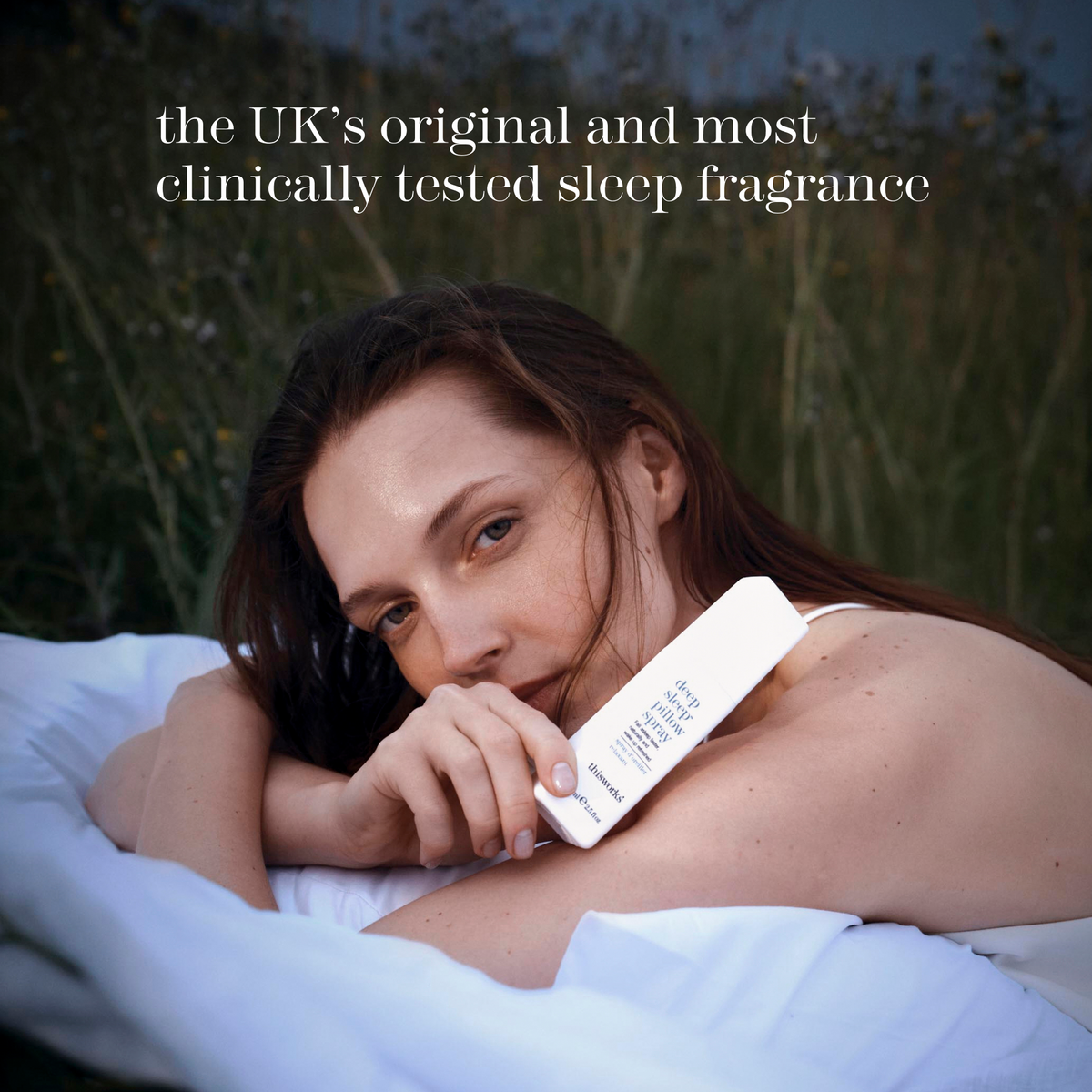 Woman lying on white bedding holding This Works Deep Sleep Pillow Spray near her face, with headline 'the UK's original and most clinically tested sleep fragrance' overlaid on a dusk meadow background.